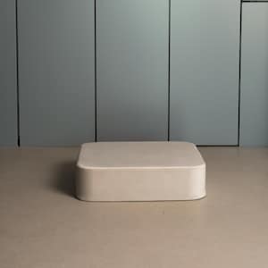 Puede incluir: Un otomán rectangular beige con bordes redondeados se encuentra sobre un suelo marrón claro. El fondo presenta una pared gris claro con líneas verticales negras y una puerta con manija y un pequeño cuadrado negro.