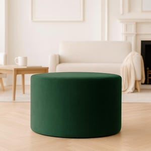 Pode incluir: Um pufe redondo verde esmeralda. A peça de mobiliário cilíndrica é o ponto focal de uma sala de estar brilhante e arejada. O pufe é feito de um material macio e aveludado. A sala possui uma lareira, um sofá e uma mesa de centro de madeira.