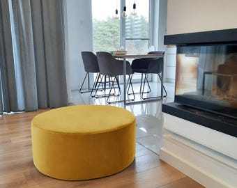 Puede incluir: Un otomán redondo de terciopelo amarillo mostaza, de unos 60 cm de diámetro, se encuentra sobre un suelo de madera. Esta pieza de mobiliario baja y cilíndrica es el punto focal de un salón moderno, con una mesa de comedor y una chimenea al fondo.