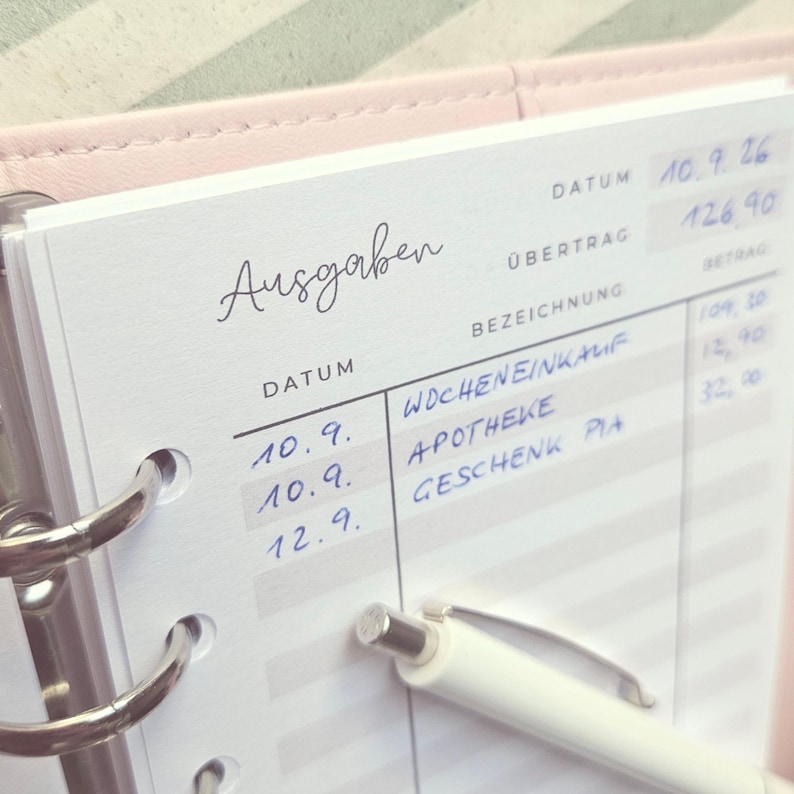 May include: A white expense tracker page with the German heading Ausgaben, featuring columns for Datum, Bezeichnung, and Betrag. The page is held in a light pink binder with silver rings and includes handwritten entries in blue ink and a white pen.