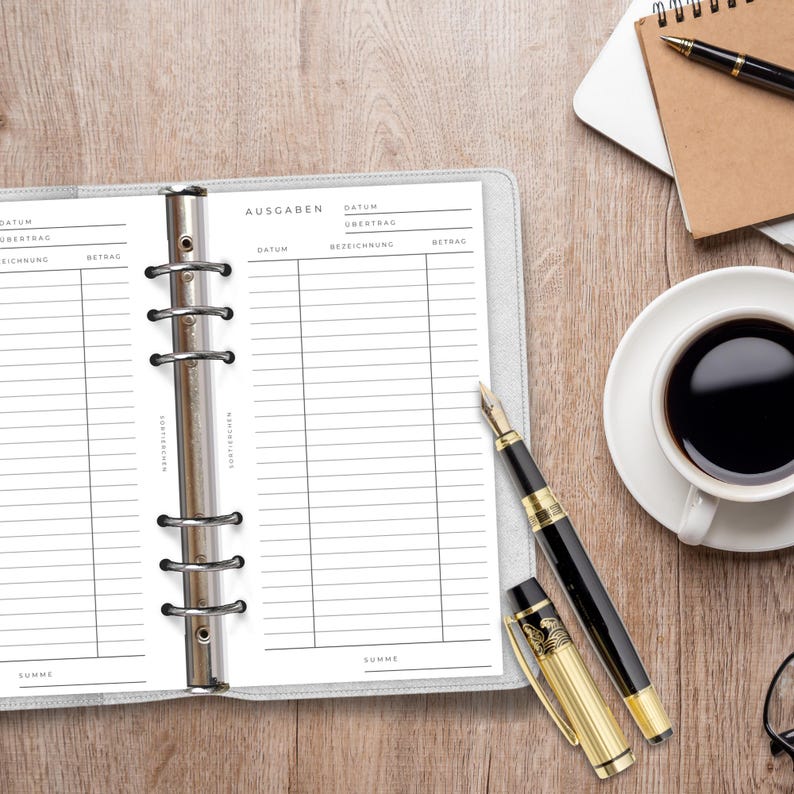 May include: A six-ring binder with white pages featuring black printed columns for tracking expenses, labeled with the German word Ausgaben. A black and gold fountain pen with its cap removed lies beside the binder, along with a cup of black coffee.