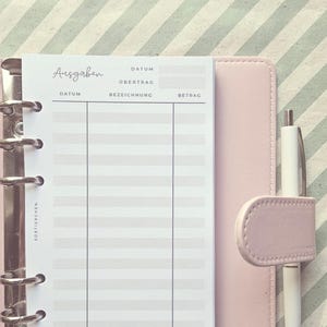 May include: A light pink six-ring binder containing a white expense tracker insert with the German text Ausgaben, Datum, &Uuml;bertrag, Bezeichnung, and Betrag printed in black. A white pen is held in the side loop of the binder cover.