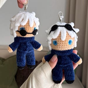 May include: Two plush crochet keychains featuring characters with white hair, bright blue eyes, and black sunglasses. Each figure wears a dark navy blue long-sleeved outfit with a high collar and black shoes, attached to a silver metal lobster clasp.