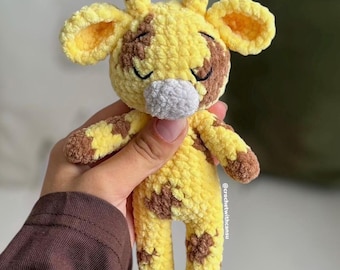 May include: A handmade crochet giraffe toy in yellow and brown. The giraffe has closed eyes, a gray nose, and brown spots. The ears and the tips of the horns are brown. The toy is held in a person's hand.