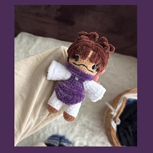 May include: A small, handmade crochet doll featuring a plush brown yarn hairstyle, a purple vest, and white long sleeves and trousers. The doll has two small black bead eyes and a simple stitched mouth, crafted from soft, textured yarn materials.