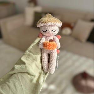 Könnte beinhalten: Handgefertigte Häkelpilzfigur in zartem Rosa mit beiger Kappe. Die Figur hält einen orangefarbenen Kürbis und hat geschlossene Augen. Ein skurriles Deko-Objekt.