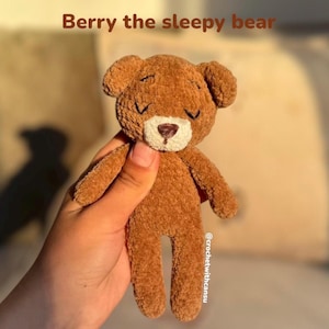 Könnte beinhalten: Ein brauner gehäkelter Bär namens "Berry the sleepy bear" mit geschlossenen Augen und einer weißen Schnauze. Der Bär wird in einer Hand gehalten und hat kleine Arme und Beine. Der Text "Berry the sleepy bear" steht über dem Bären.
