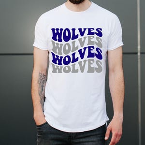 May include: White t-shirt featuring the word "WOLVES" in a layered design of blue and grey. The shirt has a crew neck and is made of a soft material. The person is wearing dark blue jeans.