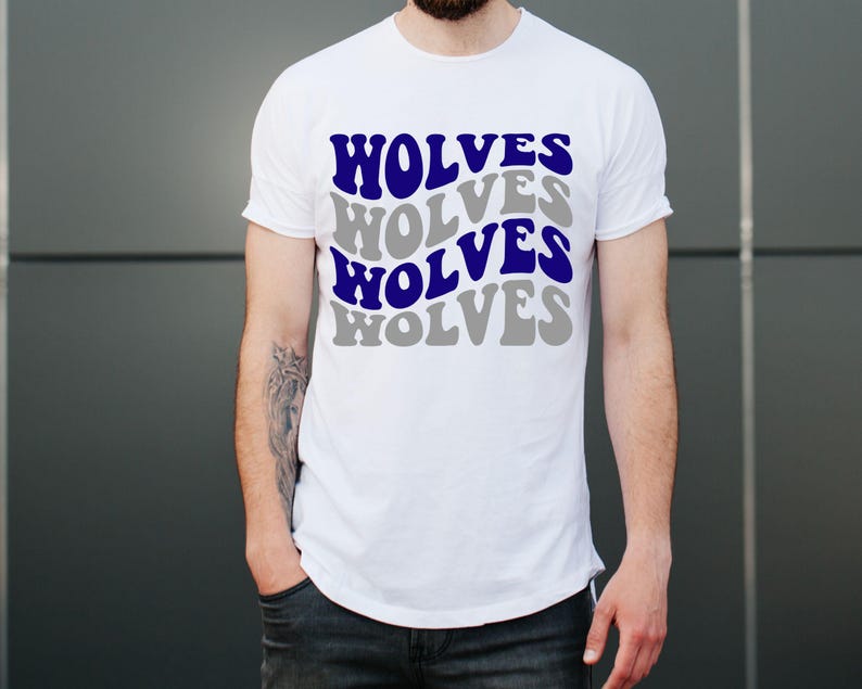 May include: White t-shirt featuring the word "WOLVES" in a layered design of blue and grey. The shirt has a crew neck and is made of a soft material. The person is wearing dark blue jeans.