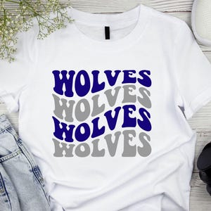 May include: White t-shirt featuring the word "WOLVES" in a repeating pattern of blue and grey text. The shirt is displayed on a wooden surface with white trainers, headphones, and a pair of jeans.