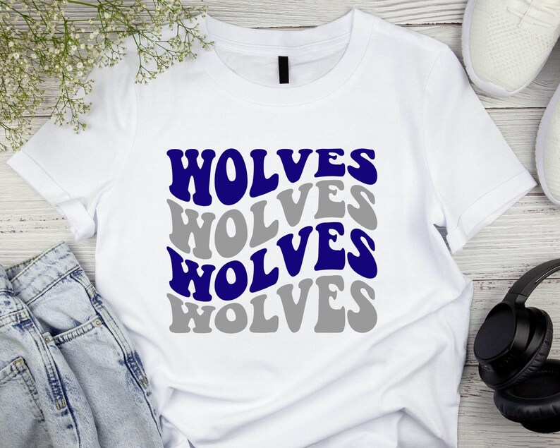 May include: White t-shirt featuring the word "WOLVES" in a repeating pattern of blue and grey text. The shirt is displayed on a wooden surface with white trainers, headphones, and a pair of jeans.
