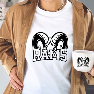 May include: A white t-shirt with a black graphic of a ram's head and the word "RAMS" in a bold font. A white mug with a black graphic of a ram's head and the word "RAMS" in a bold font is being held by a person wearing a brown jacket.