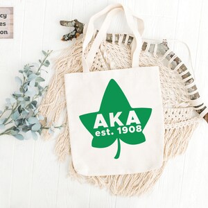 May include: A white canvas tote bag featuring a green ivy leaf design with the text "AKA est. 1908" in white.