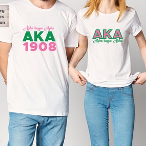 May include: Two white t-shirts with the letters AKA in pink and green. The larger shirt has the text "Alpha kappa Alpha" and "1908" below the letters. The smaller shirt has the text "Alpha kappa Alpha" below the letters.