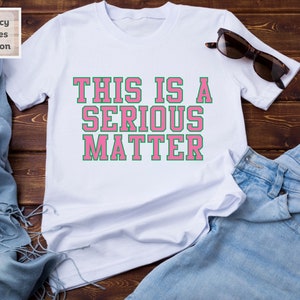 May include: White t-shirt with the text "THIS IS A SERIOUS MATTER" in pink letters with a green outline.