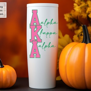 May include: White tumbler with a pink and green "AKA" design. The letters are outlined in green and the words "Alpha Kappa Alpha" are written in cursive green lettering.