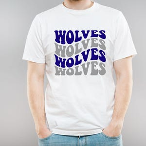 May include: White t-shirt featuring the word "WOLVES" in a layered design of blue and grey text. The shirt is worn by a person, with their hands in their pockets. The t-shirt is made of a soft material.