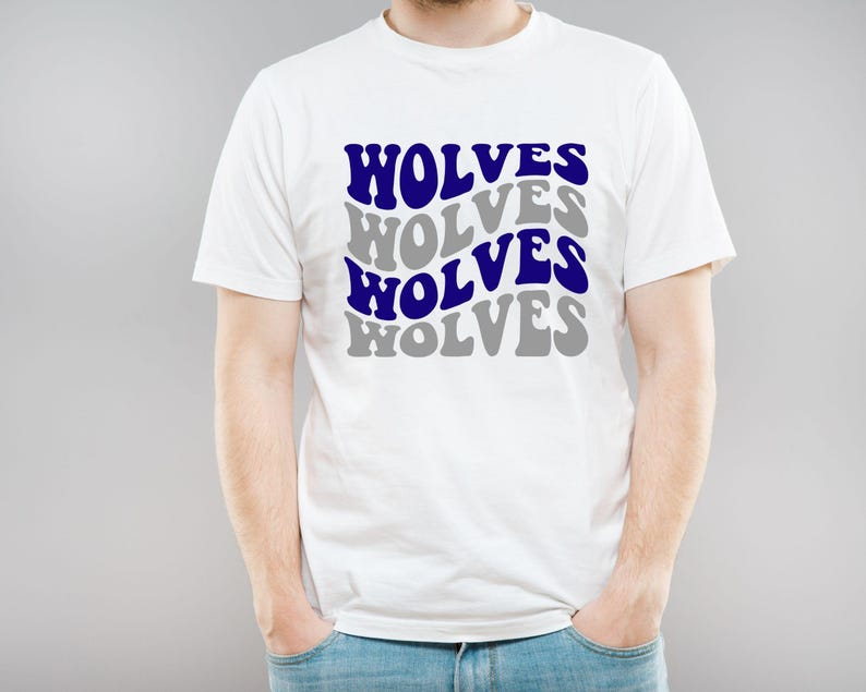 May include: White t-shirt featuring the word "WOLVES" in a layered design of blue and grey text. The shirt is worn by a person, with their hands in their pockets. The t-shirt is made of a soft material.