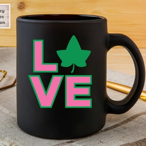 May include: Black ceramic coffee mug with a pink and green "LOVE" design featuring a green ivy leaf.