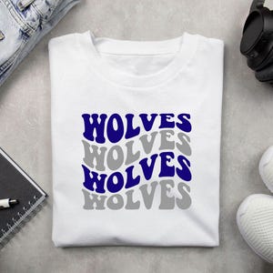 May include: White t-shirt featuring the word "WOLVES" in a stacked design. The word is in a retro font, alternating between blue and grey. The shirt is displayed on a flat surface with a notebook, pen, headphones, and trainers.