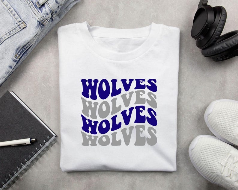 May include: White t-shirt featuring the word "WOLVES" in a stacked design. The word is in a retro font, alternating between blue and grey. The shirt is displayed on a flat surface with a notebook, pen, headphones, and trainers.