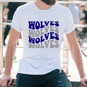 May include: White t-shirt featuring the word "WOLVES" in a repeating, wavy pattern. The text is in blue and grey, giving a retro layered look. A casual, everyday wear item.