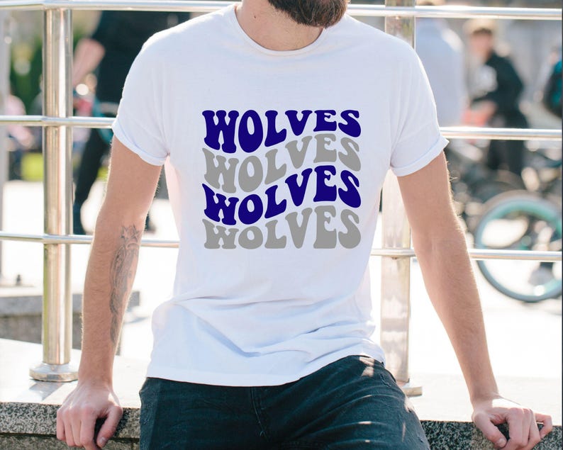 May include: White t-shirt featuring the word "WOLVES" in a repeating, wavy pattern. The text is in blue and grey, giving a retro layered look. A casual, everyday wear item.