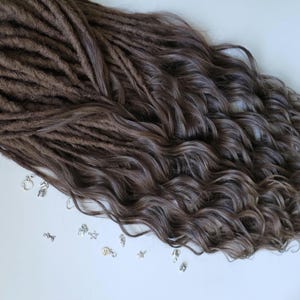 May include: Brown dreadlocks with wavy ends and small silver charms scattered on a white surface. The dreadlocks are a medium brown color, transitioning to a lighter shade at the ends. The charms include stars, keys, and other small shapes.