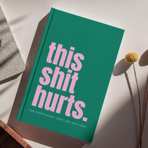 May include: A green hardcover journal with pink text that reads this shit hurts. for everything i can't say out loud. The book is positioned next to two dried yellow billy ball flowers on thin stems, creating a simple and clean visual composition.