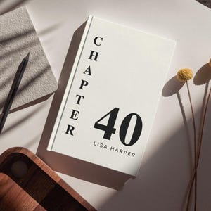 Peut inclure: Un livre à couverture rigide blanc intitulé Chapter 40 par Lisa Harper avec un texte noir en gras sur la couverture. Le livre est placé à côté d'un crayon noir, d'un bloc-notes gris texturé et de deux fleurs jaunes séchées sur une surface blanche.