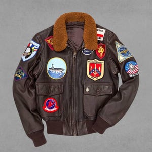 May include: A brown leather bomber jacket with a shearling collar and multiple patches. The patches include a ship, a trident, a plane, and a cat. The jacket has a zipper closure and two pockets.