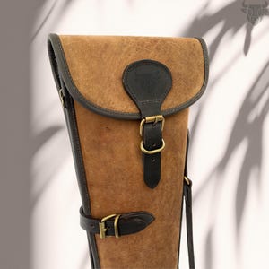 Puede incluir: Una funda de arma de cuero marrón con una correa y una hebilla de cuero negro. La funda tiene una solapa con un logotipo de cabeza de toro de cuero negro.