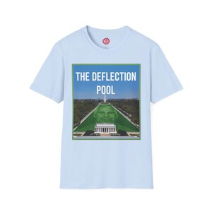 May include: A light blue short-sleeve t-shirt featuring a rectangular graphic print of the Lincoln Memorial Reflecting Pool with a green face superimposed on the water. The text THE DEFLECTION POOL is printed in white capital letters above the image.
