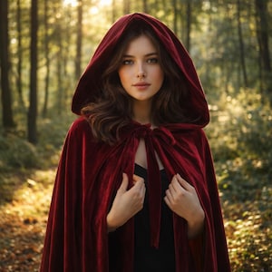 May include: A woman wearing a burgundy velvet hooded cloak, tied at the neck. The cloak is a deep red color and drapes over a black top. The background is a blurred forest scene with sunlight filtering through the trees.