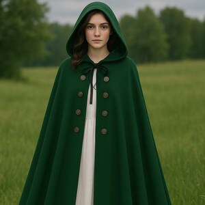 Puede incluir: Una capa con capucha verde oscuro con seis botones decorativos en la parte delantera. La capa está hecha de un material grueso y tiene una capucha grande. Una cinta negra está atada al cuello. La persona lleva un vestido blanco.