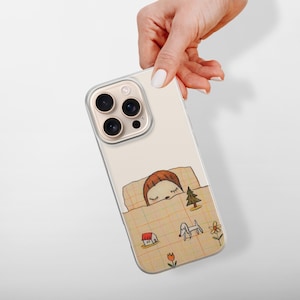 Op de afbeelding: Een beige iPhone-hoesje met een grillige illustratie van een persoon die onder een deken slaapt. Het ontwerp bevat een huis, een hond, een boom en bloemen. De hoes heeft een transparante rand en een camerabult.