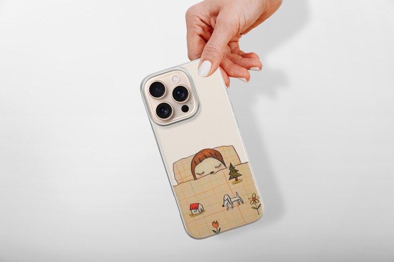 Op de afbeelding: Een beige iPhone-hoesje met een grillige illustratie van een persoon die onder een deken slaapt. Het ontwerp bevat een huis, een hond, een boom en bloemen. De hoes heeft een transparante rand en een camerabult.