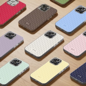 Puede incluir: Una colección de fundas para teléfonos en varios colores, cada una con un patrón de lunares y iniciales personalizadas. Los colores incluyen rojo, marrón, azul claro, lavanda, blanco, rosa, amarillo, verde y azul marino. Las fundas están dispuestas sobre una superficie de madera.