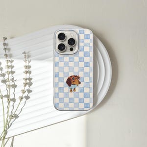 May include: A phone case with a light blue and white checkered pattern. The case features a watercolor illustration of a dachshund wearing sunglasses and a blue sweater. The phone case is resting on a white plate.