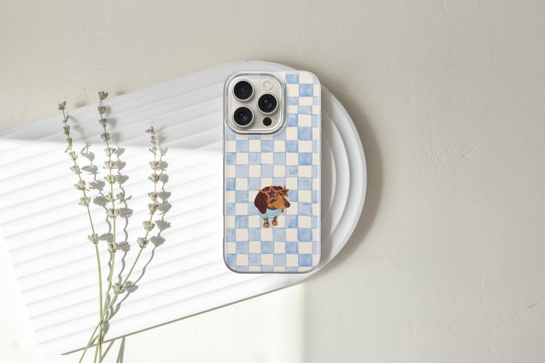 May include: A phone case with a light blue and white checkered pattern. The case features a watercolor illustration of a dachshund wearing sunglasses and a blue sweater. The phone case is resting on a white plate.