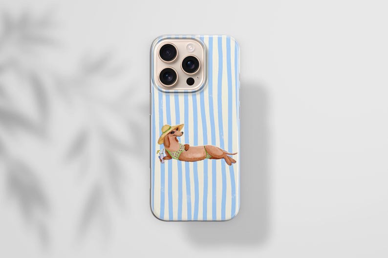 May include: A phone case with a light blue and white striped pattern. The case features an illustration of a dachshund wearing a sun hat and swimsuit, lounging with a drink. The phone case has a camera bump with three lenses.