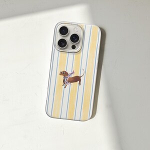 May include: A smartphone case with a yellow and white striped pattern. A brown dachshund dog illustration with a scarf is centered on the case. The case has a clear border around the camera lenses.