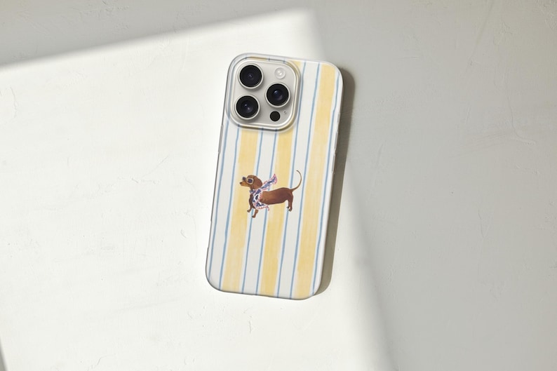 May include: A smartphone case with a yellow and white striped pattern. A brown dachshund dog illustration with a scarf is centered on the case. The case has a clear border around the camera lenses.