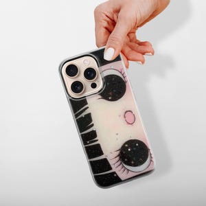 Może przedstawiać: Etui na telefon z fantazyjnym wzorem przedstawiającym twarz z dużymi, gwieździstymi oczami i rzęsami. Etui ma czarno-biały wz&oacute;r z boku i złotą osłonę obiektywu aparatu.