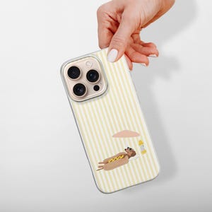 May include: A clear phone case with a yellow and white striped pattern. The case features a cartoon illustration of a hot dog relaxing under a beach umbrella. The phone case is held by a hand.