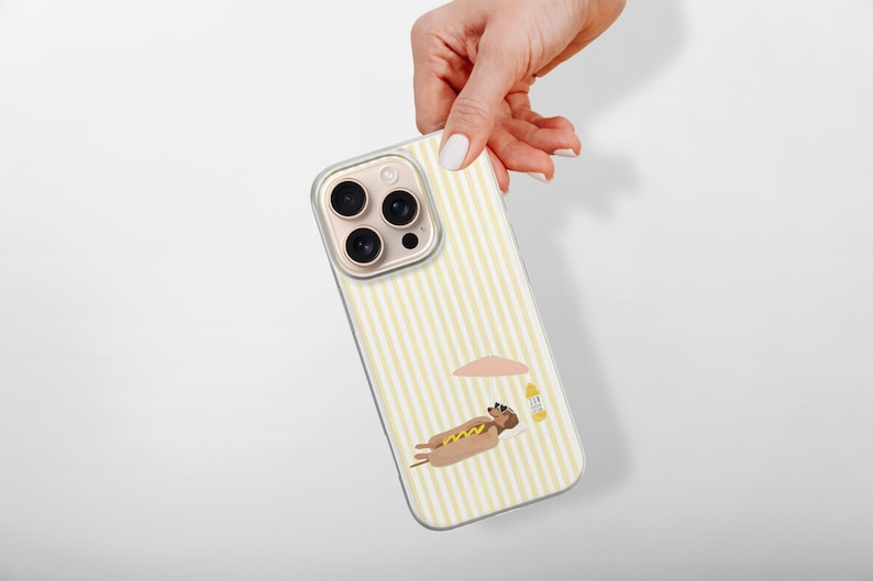 May include: A clear phone case with a yellow and white striped pattern. The case features a cartoon illustration of a hot dog relaxing under a beach umbrella. The phone case is held by a hand.