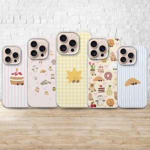 Puede incluir: Cinco fundas para teléfono con varios diseños. Las fundas presentan ilustraciones de un pastel, un sol y otros personajes lindos. Las fundas tienen un protector de lente de cámara transparente y están dispuestas sobre una superficie de madera.