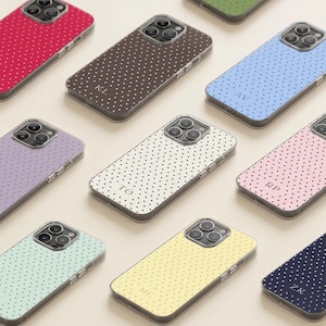 Op de afbeelding: Een collectie smartphonehoesjes in diverse effen kleuren met witte of zwarte stippenpatronen. Verschillende hoesjes tonen monogrammen van twee letters zoals KL, AV, TO, RP, MC en ZK. Elk hoesje heeft een uitsparing voor een triple-camerasysteem.