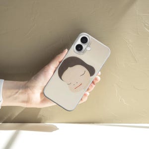 Op de afbeelding: Een beige smartphonehoesje met een minimalistische illustratie van een gezicht met gesloten ogen en bruin haar. De hoes wordt in een hand gehouden, met een wit horloge om de pols. De achtergrond is een getextureerd, lichtbruin oppervlak.