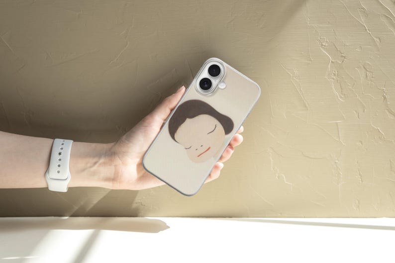 Op de afbeelding: Een beige smartphonehoesje met een minimalistische illustratie van een gezicht met gesloten ogen en bruin haar. De hoes wordt in een hand gehouden, met een wit horloge om de pols. De achtergrond is een getextureerd, lichtbruin oppervlak.
